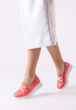 Jambes portant une jupe blanche et des mocassins rose corail avec des semelles blanches et des lacets décoratifs blancs, marchant sur un fond clair uni.
