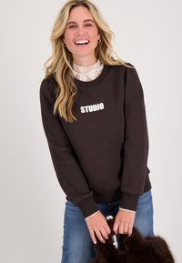 Bruine sweatshirt met de tekst "STUDIO", geribbelde manchetten en een relaxed fit. Gelaagd over een witte kant-top, gecombineerd met blauwe jeans.