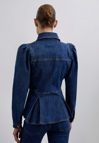 Mørkeblå denimjakke med puffede ærmer, en tætsiddende talje og peplum detaljer bagpå. Syede sømme og en gylden knapdetalje på ærmet.