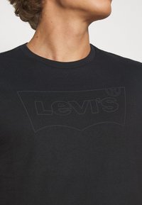 Detailní záběr osoby nosící černé tričko Levi's s jemným, vyraženým logem Levi's na hrudi.