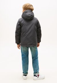 Giacca grigia imbottita con cappuccio, vista da dietro. Indossata con jeans azzurri chiari e sneaker bianche. Sfondo semplice.