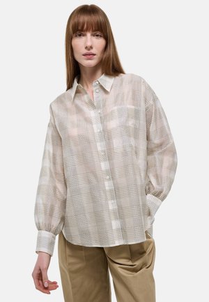 Femme portant une chemise ample à carreaux beige et blanche à manches longues et un pantalon beige sur fond uni.