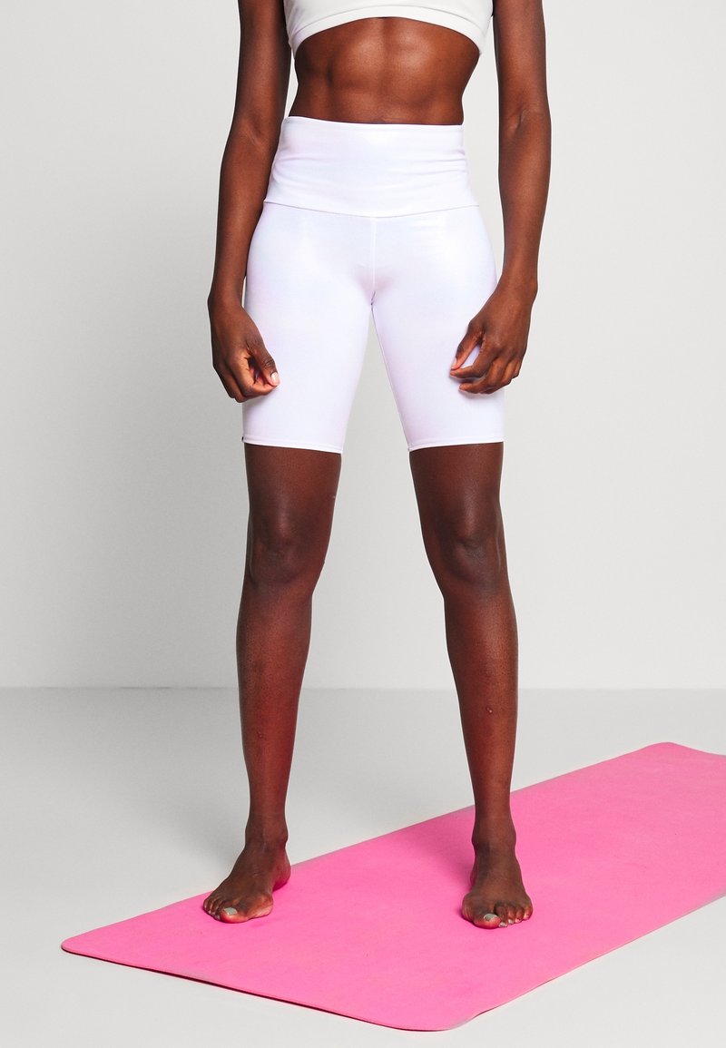 White high-waisted bike shorts made of stretchy material, fitted design, paired with a matching cropped top, standing on a pink mat.