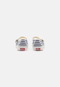 Sapatilhas slip-on de canvas com padrão listrado em branco e azul, solas de borracha e detalhe em fivela de metal. Logotipo vermelho na parte de trás.