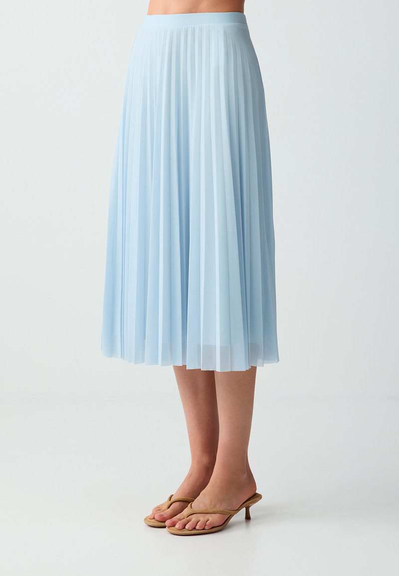 Jupe midi plissée bleu clair portée avec des sandales à talons couleur beige, montrée de la taille aux pieds sur un fond uni.