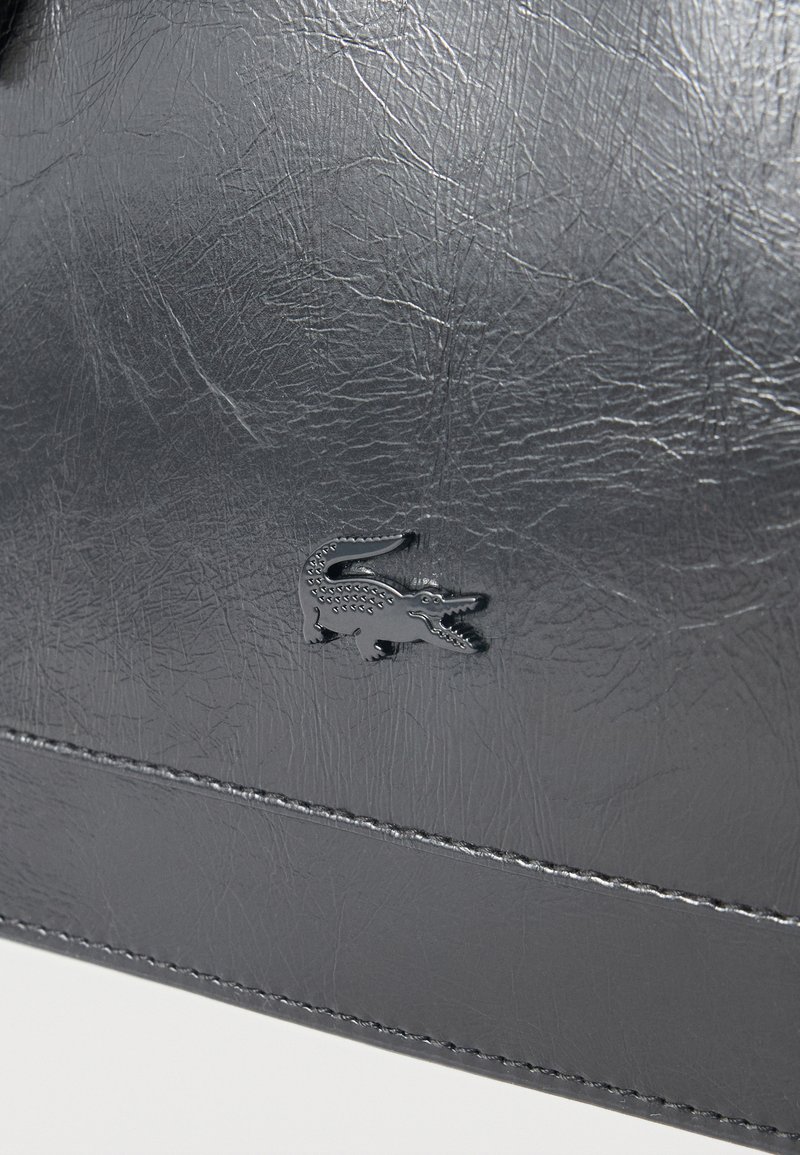 Superficie in pelle nera con piccolo logo coccodrillo in rilievo e cuciture visibili vicino al bordo inferiore.