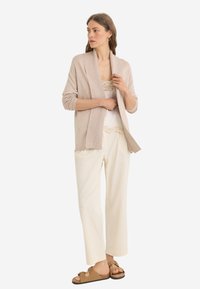 Cardigan beige con colletto scialle, indossato su una maglietta bianca con bordi in pizzo e pantaloni crema; completato con sandali marroni senza fibbie.