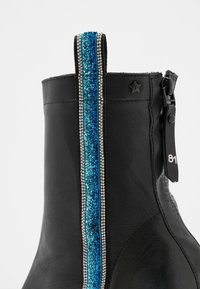 Bottine en cuir noir avec une bande verticale à paillettes bleues bordée de perles argentées, un emblème en forme d'étoile, et une fermeture éclair latérale avec un tirant "81".