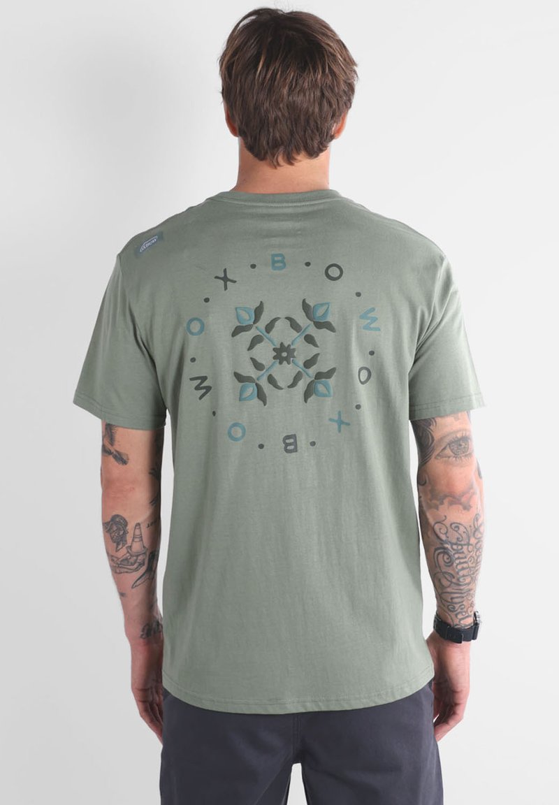 T-shirt vert olive à manches courtes, présentant un motif floral circulaire en bleu et noir au dos, fabriqué en tissu léger.