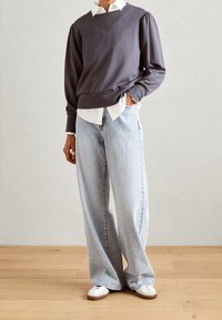 Grijze pullover sweatshirt met poffende mouwen, gelaagd over een wit overhemd met kraag. Gepaard met lichtblauwe wijde jeans en witte sneakers.