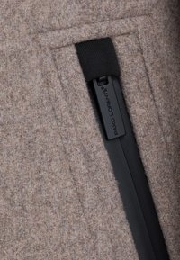 Brązowy filcowy materiał o gładkiej fakturze, z czarnym zamkiem błyskawicznym i czarną metką z logo, zintegrowany w funkcjonalnym designie.