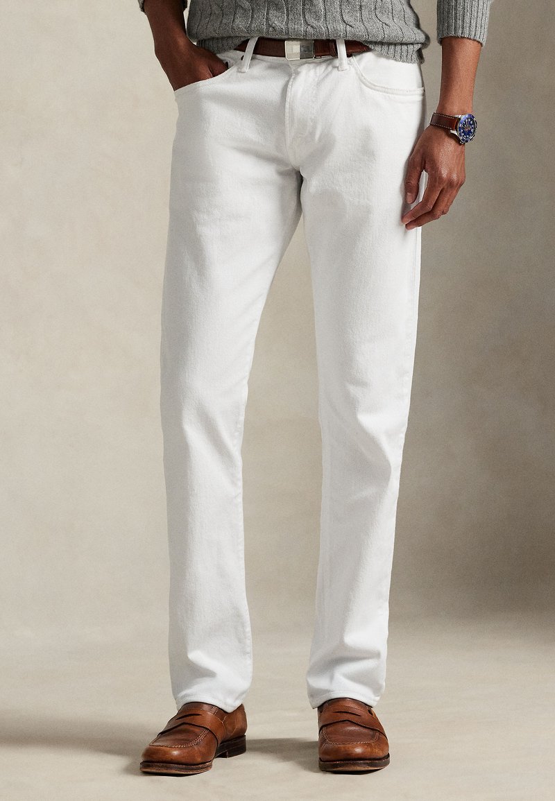 Calças brancas de algodão com corte direito, com cinco bolsos e cinto. Usadas com sapatos de couro castanho e um suéter cinza.