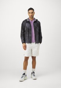 Lättviktig, svart mönstrad jacka med huva, bärs över en lila polo-skjorta. Vita shorts och sportiga sneakers kompletterar klädseln.