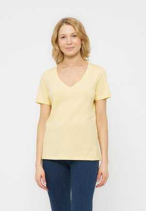 Γυναίκα με ξανθά μαλλιά που φοράει ανοιχτό κίτρινο μπλουζάκι με V και κοντά μανίκια, καθώς και σκούρο μπλε τζιν, στέκεται μπροστά σε λευκό φόντο.