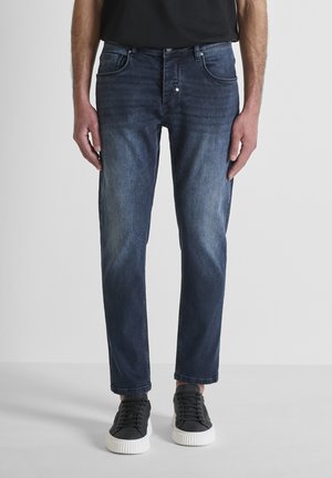 Uomo che indossa jeans slim fit a lavaggio scuro e sneakers nere con suole bianche, in piedi davanti a uno sfondo chiaro e uniforme.