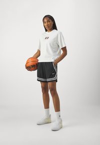 White short-sleeve t-shirt with logo, paired with black basketball shorts featuring white stripes, and white sneakers with accents. Holding a basketball.