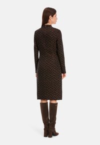 Langärmliges Kleid in Dunkelbraun mit Zickzackmuster, Seitenschlitz und taillierter Silhouette. Kombiniert mit knielangen braunen Wildlederstiefeln.