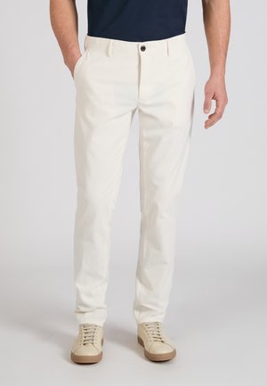 Mężczyzna ubrany w białe, dopasowane spodnie i beżowe trampki, stojący z jedną ręką w kieszeni na jednolitym tle.