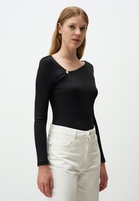 Zwarte lange mouw top van ribmateriaal met een ronde halslijn en een decoratieve zilveren lus accent bij de schouder. Gekleed met witte broek.