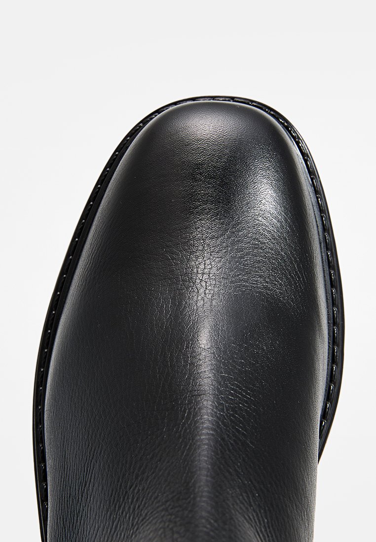 Cap de botte en cuir noir avec une texture lisse, une forme arrondie et des coutures contrastées le long du bord, présentant un éclat subtil.