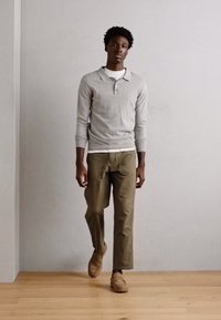 Grå langærmet polo med krave lagd over en hvid skjorte, parret med olivengrønne bukser og brune slip-on sko, sat mod en lys baggrund.
