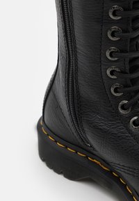 Svart läderankelboot med sidodragkedja, metallöglor, gul söm på sulan och texturerad yta.