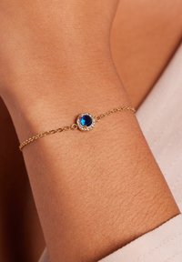 Gouden kettingarmband met een ronde blauwe edelsteen als middelpunt, omringd door een cirkel van heldere stenen, op een gebruinde pols.