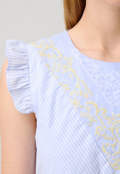 Blusa senza maniche azzurro chiaro con righe bianche, dettaglio arricciato sulla spalla e motivo ricamato giallo e bianco vicino allo scollo.