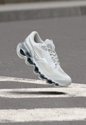 Chaussure de sport blanche et grise avec une semelle incurvée unique, en l'air au-dessus de l'asphalte avec des marquages blancs peints et de petits débris éparpillés.