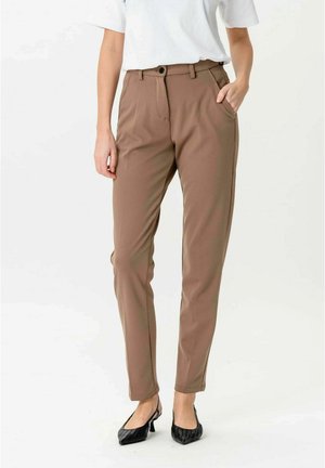 Person iført brune slim-fit bukser, hvid kortærmet top og sorte spidse hæle, stående med den ene hånd i lommen.