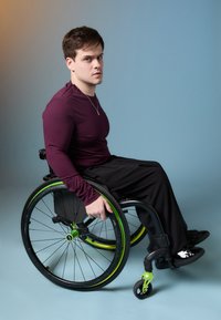 Zöld díszítésű, fekete kerekű tolószék, amelyben egy maroon hosszú ujjú pólót és fekete nadrágot viselő személy ül, jobbra nézve.