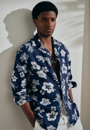 Jeune homme portant une chemise bleu marine à imprimé floral et léopard avec les manches retroussées, un pantalon blanc et un collier de perles, debout contre un mur clair.