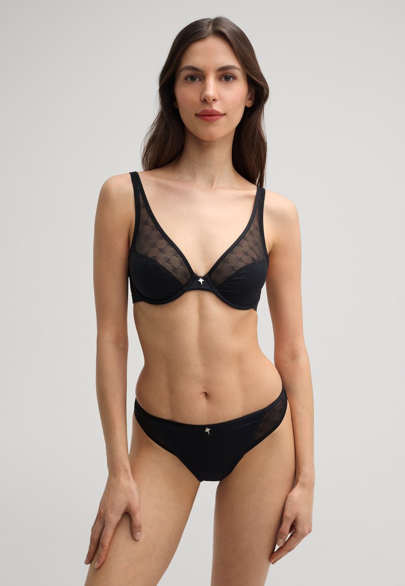 Zwart lingerie set bestaande uit een plunge bh met transparante panelen en een bijpassende slip, versierd met delicate stiksels en een klein embleem.
