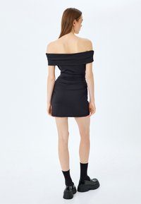 Sort off-shoulder kjole med rynkedetaljer, tætsiddende silhuet, kort længde, og parret med kraftige sorte sko og sokker.