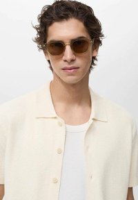Occhiali da sole con una montatura beige traslucida e lenti marroni scure, indossati da una persona in una camicia button-up crema a trama sopra un top bianco.
