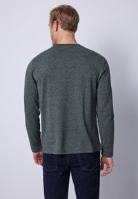 Graues Langarmshirt aus strukturiertem Stoff, mit Rundhalsausschnitt und seitlichen Schlitzen, kombiniert mit dunklen Denim-Jeans. Rückansicht gezeigt.