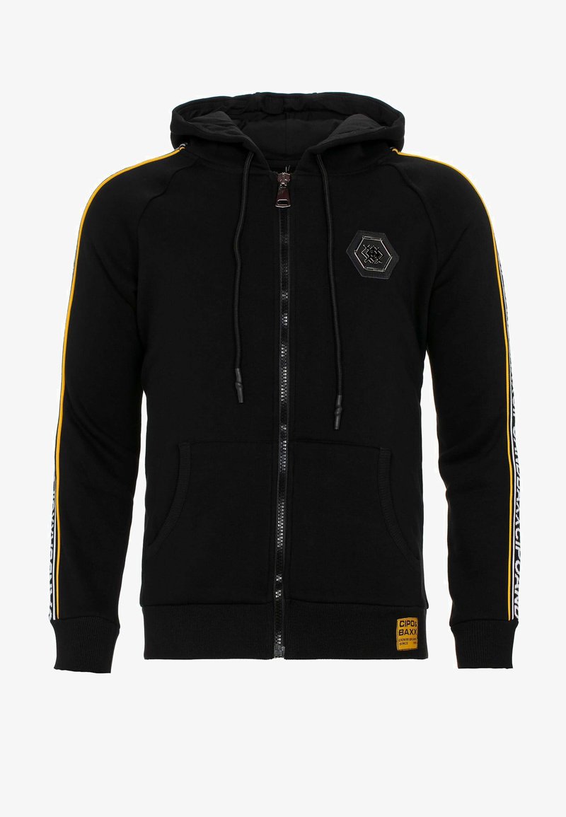 Schwarze Zip-Hoodie mit doppellagiger Kapuze, gelben Streifen an den Ärmeln, Fronttaschen und einem hexagonalen Logo-Patch auf der Brust.