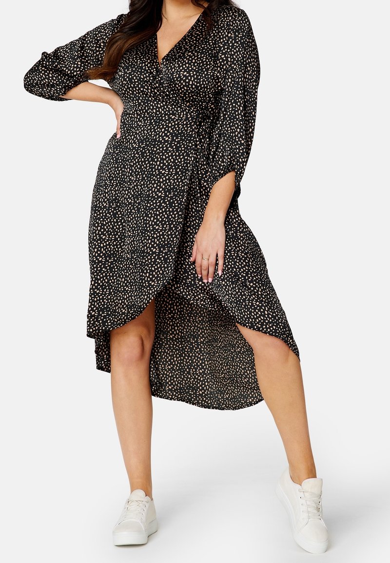 Schwarzes Wickelkleid mit weißem Punktmuster, langärmlig und mit asymmetrischem Saum, kombiniert mit weißen Sneakers. Glatte Textur, leichtes Material.
