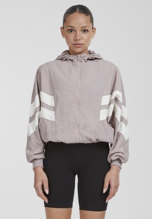 Žena s vlasmi zopnutými dozadu nosí svetlo fialovú bundu na zips s bielymi chevronovými pruhmi na rukávoch a čierne cyklistické šortky.