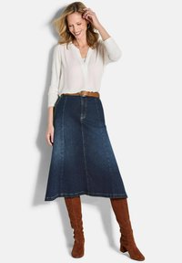 Gonna in denim a forma di A di colore blu scuro con dettagli di cucitura visibili, abbinata a una blusa leggera a maniche lunghe e una cintura e stivali in pelle marrone.