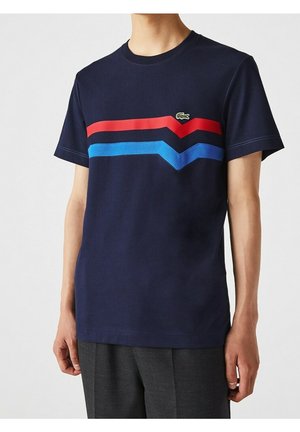 Homme portant un t-shirt bleu marine avec des bandes en chevron rouges et bleues et un petit logo de crocodile vert sur la poitrine.