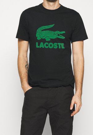 T-shirt en coton noir avec un grand logo de crocodile vert et le texte "LACOSTE" dans la même couleur. Manches courtes, col rond.