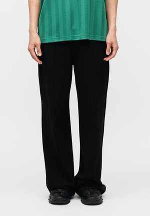 Personne portant un pantalon noir ample, des baskets noires et une chemise rayée verte, debout devant un fond uni.