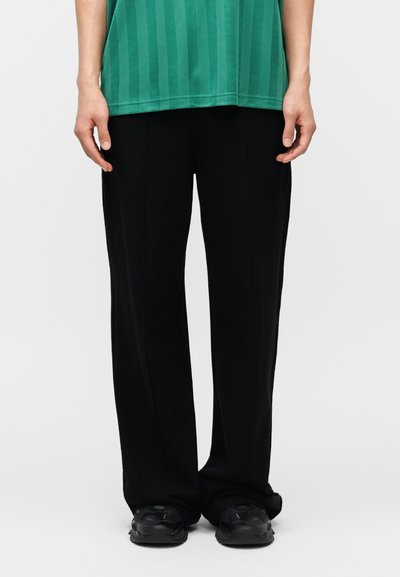 Persona vestida con pantalones negros holgados, zapatillas negras y una camisa de rayas verdes, de pie contra un fondo liso.