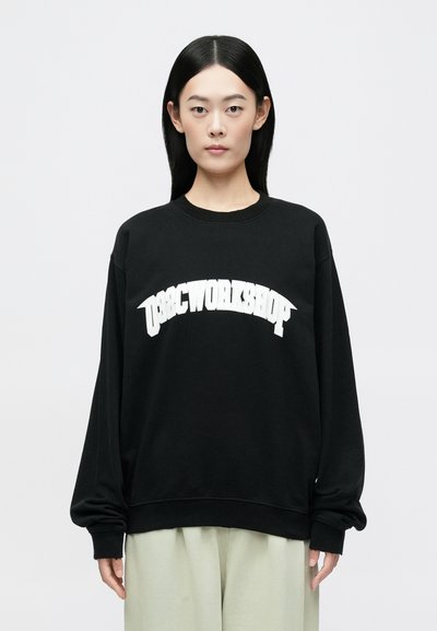 Μαύρη φόρμα με oversized εφαρμογή, που διαθέτει λευκή καμπύλη γραφιστική "USGCWORKSHOP" στο μπροστινό μέρος. Μαλακό ύφασμα, ribbed μανσέτες και στρίφωμα.