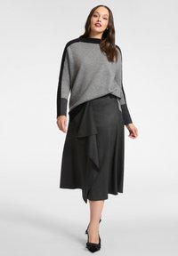 Grauer Strickpullover mit schwarzen Akzenten, kombiniert mit einem dunkelgrauen Rock mit drapiertem Detail. Schwarze Pumps vervollständigen das Outfit.