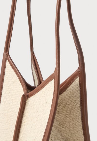 Μπεζ υφασμάτινη τσάντα ώμου με καφέ δερμάτινες λεπτομέρειες και μακριές δερμάτινες λαβές, εμφανίζεται σε κοντινό πλάνο σε ανοιχτό φόντο.