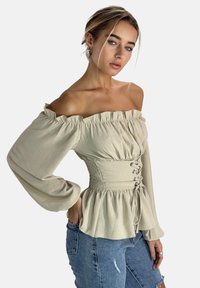 Schulterfreies Oberteil mit langen Ärmeln aus leichtem beigen Baumwollmix; geraffte Taille mit Schnürdetails; lockerer Peplum-Saum; kombiniert mit zerrissenen Jeans-Shorts.