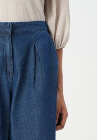 Calças de ganga com um corte descontraído, na cor azul escuro, apresentando pregas à frente e uma blusa texturizada, em bege claro, com mangas bufantes.