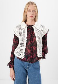 Gilet all'uncinetto aperto davanti in bianco sporco con bordi a scallop, indossato sopra una blusa floreale nera trasparente. Presenta un colletto e chiusura con bottone.